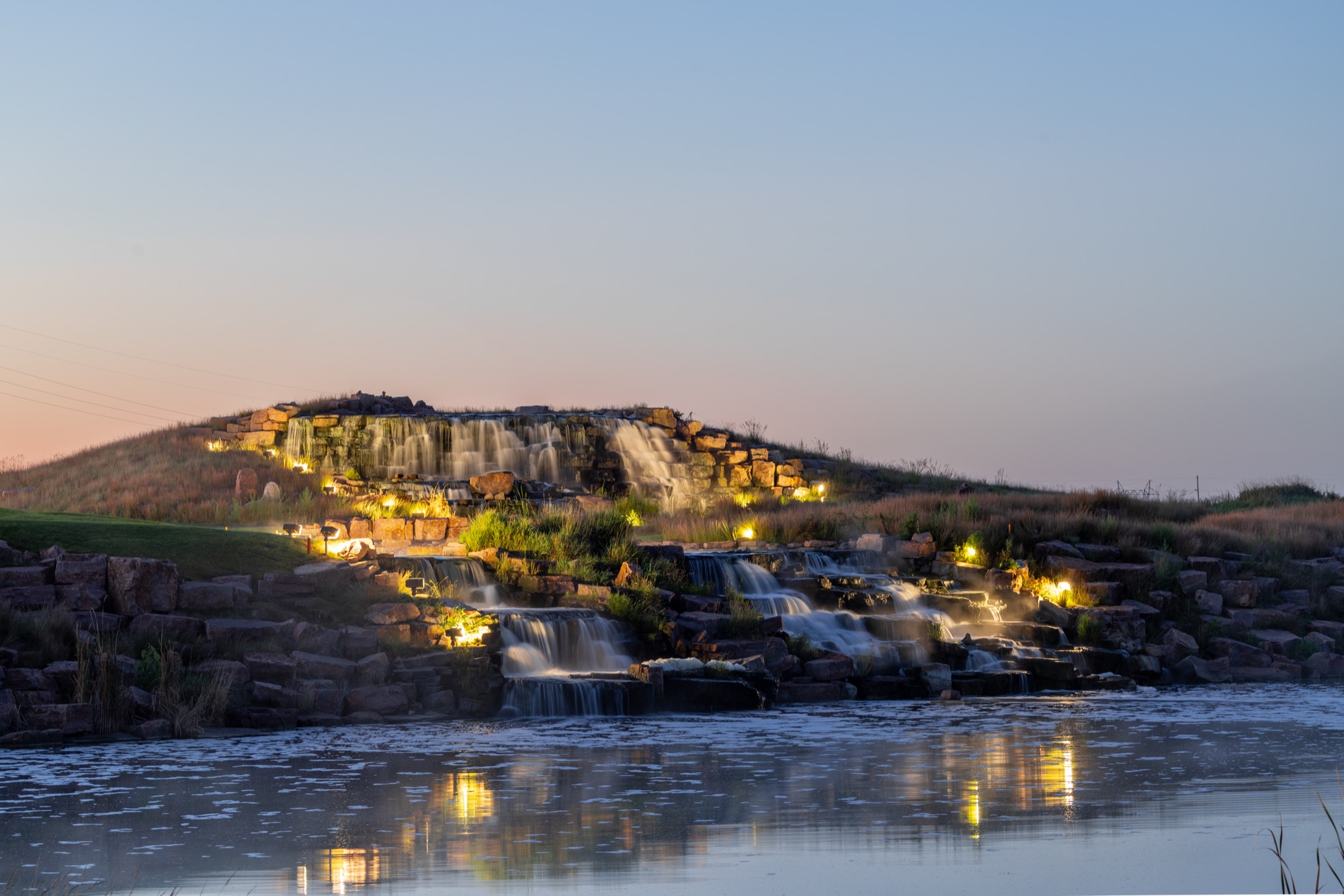 Illuminated multi-tier stone waterfall at dusk reflected in the pond below